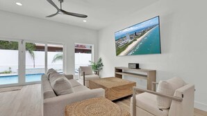 A modern living room with a large TV, comfortable seating, and a view of a pool and beach. - Pearl House - Heated Pool & Hot Tub, Sleeps 20! (South Padre Island)