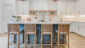 A kitchen with a central island, four wicker chairs, white cabinets, and two pendant lights. - Pearl House - Heated Pool & Hot Tub, Sleeps 20! (South Padre Island)