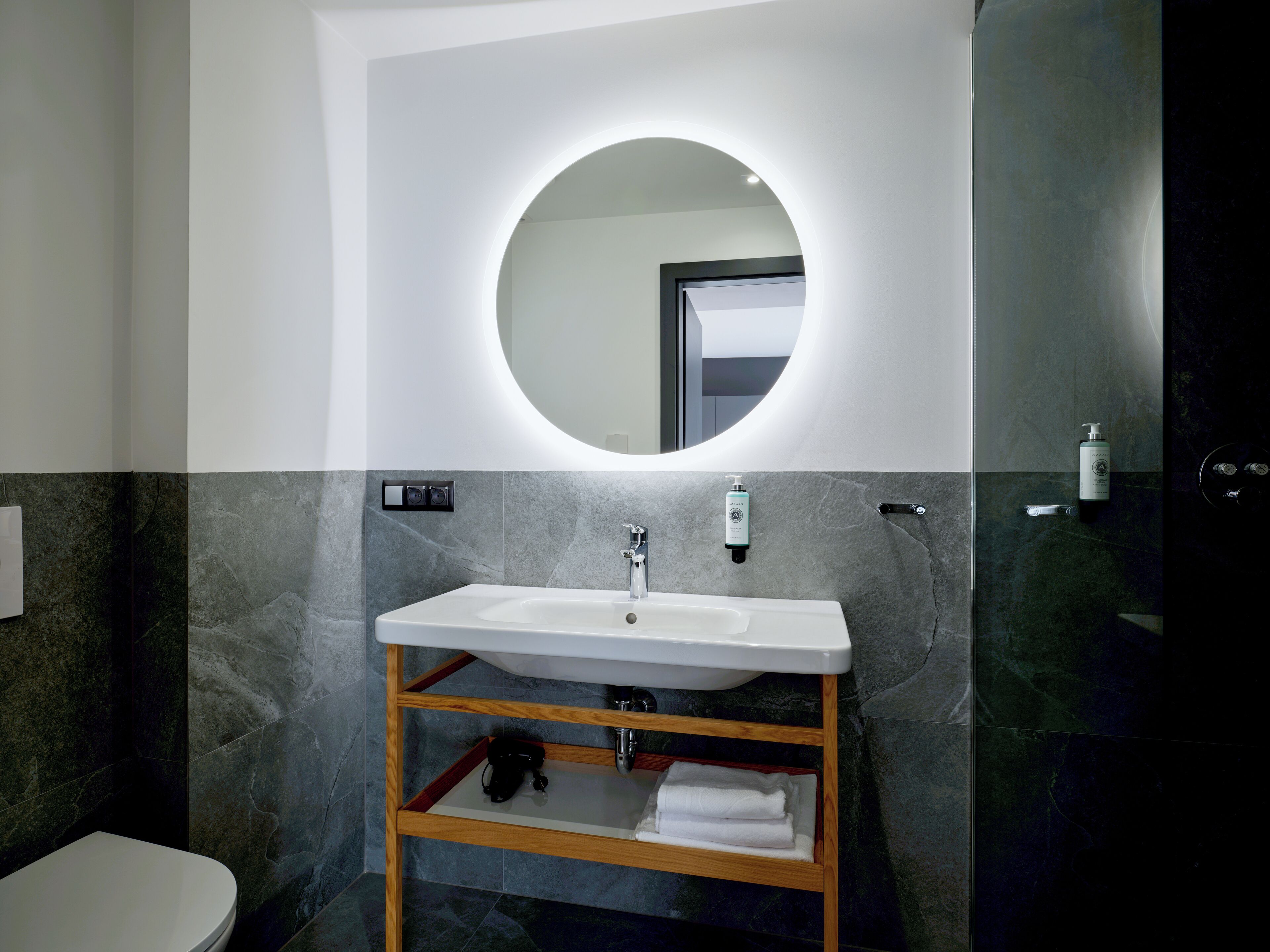 Un baño moderno con un lavamanos blanco, un mueble de tocador de madera, un espejo redondo y una pared revestida de baldosas oscuras.