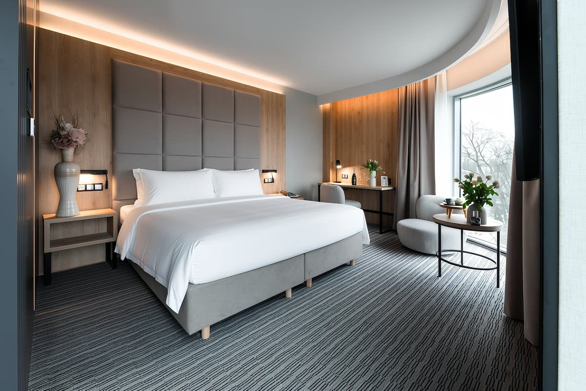 A modern hotel room with a large bed, a bedside table with a lamp, a small round table with a plant, and a view of the outdoors through a large window.