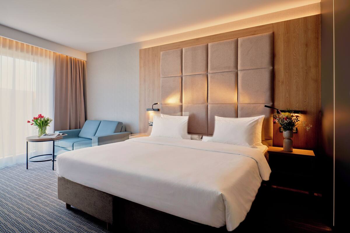 A hotel room with a large bed, a sofa, a small table with a vase of flowers, and a nightstand with a lamp.