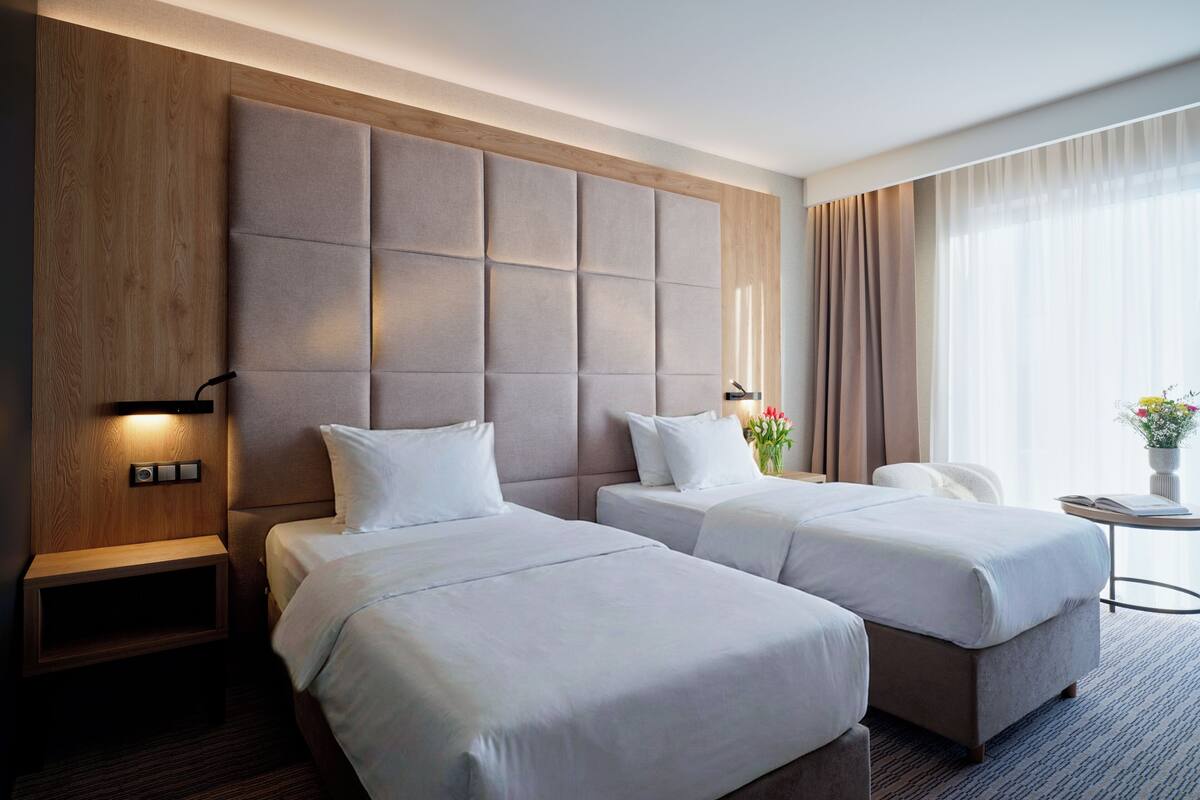 A hotel room with two beds, a wooden accent wall, a nightstand with a lamp, and a view of the outdoors through sheer curtains.