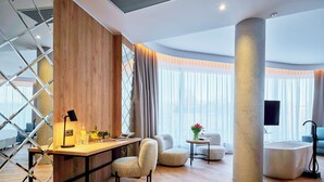 A modern hotel room with a wooden desk, two armchairs, a small round table, a large window with curtains, and a bathtub.