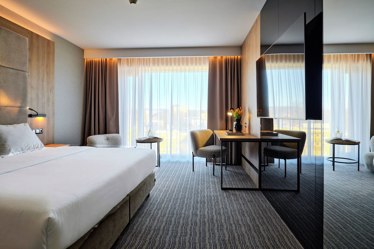 A modern hotel room with a large bed, a desk with chairs, a TV, and a view of the city.