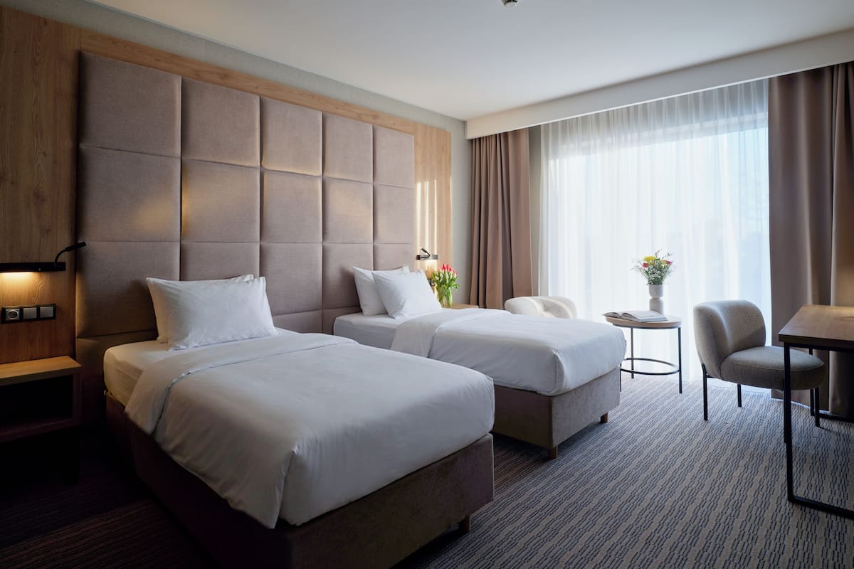 A hotel room with two beds, a small table with a chair, a desk with a chair, and a large window with curtains.