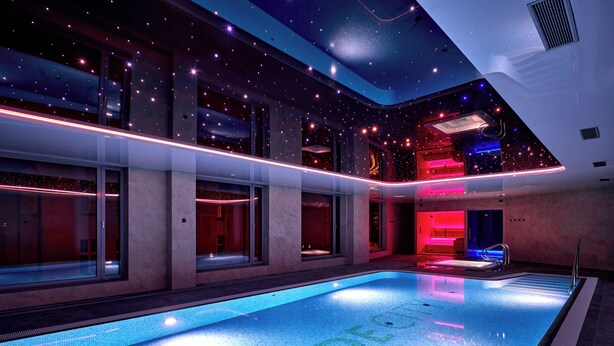 A modern indoor swimming pool with clear water, surrounded by glass walls and illuminated by ambient lighting.