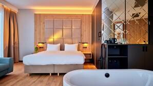 A modern hotel room with a large bed, a sitting area, a bathtub, and a kitchen area.