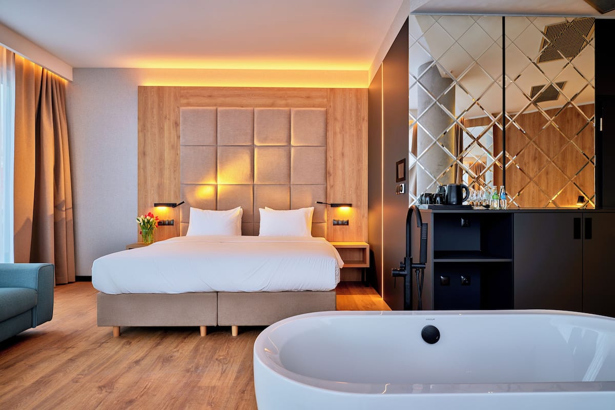 A modern hotel room with a large bed, a sitting area, a bathtub, and a kitchen area.