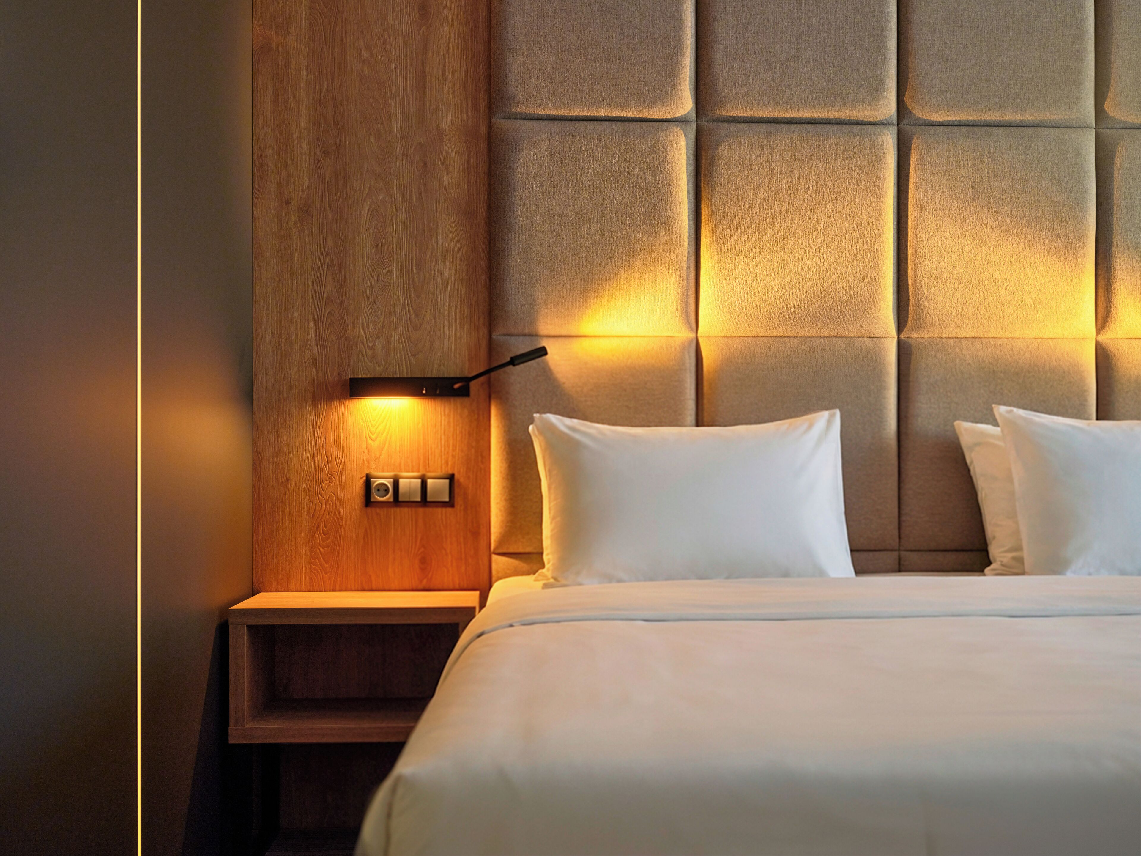 Una habitación de hotel con una mesita de noche de madera, una lámpara de pared y una cama con ropa blanca.