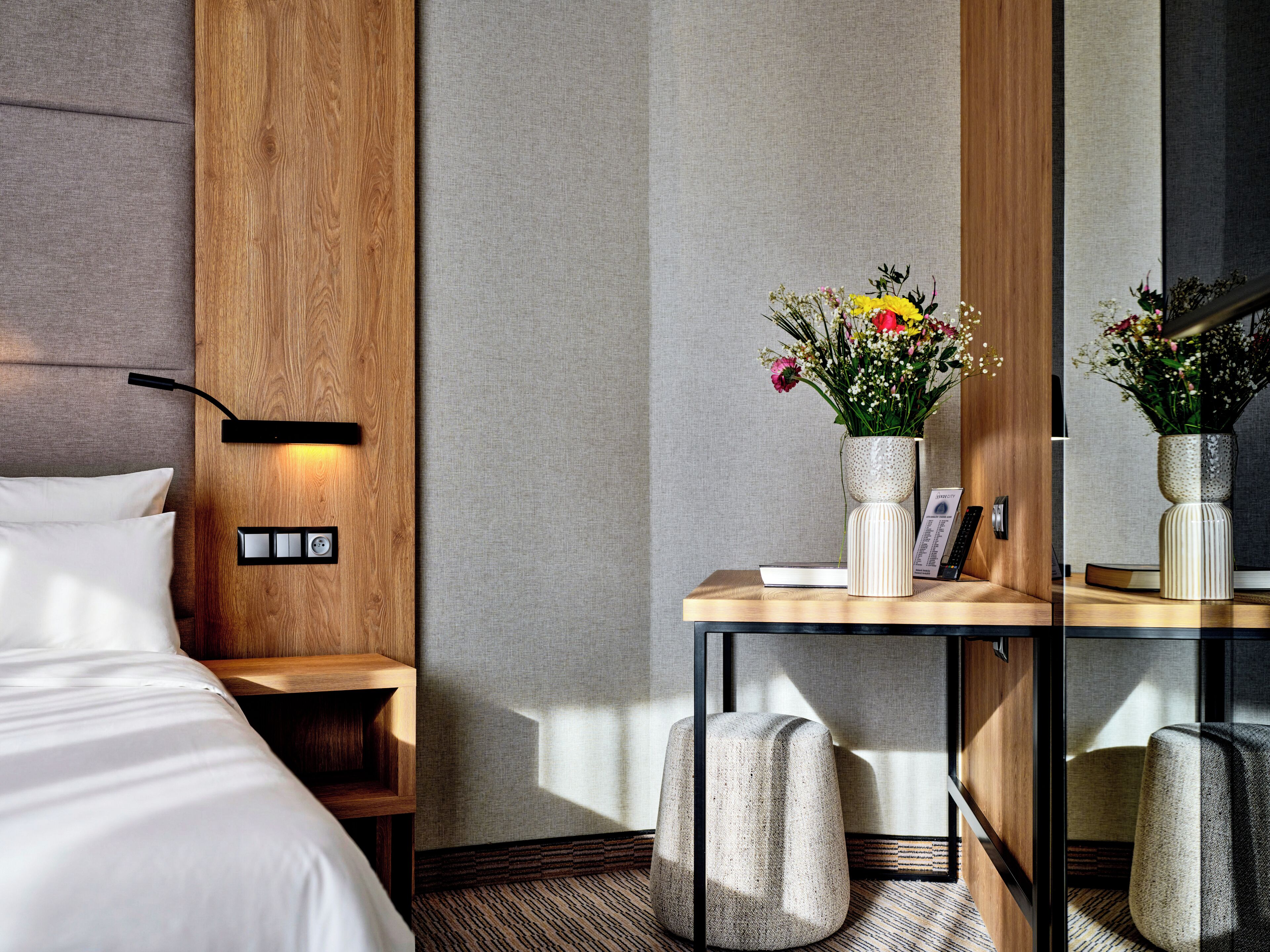 Una habitación de hotel moderna con cabecera de madera, una mesita de noche con un florero, un espejo y una cama con ropa blanca.