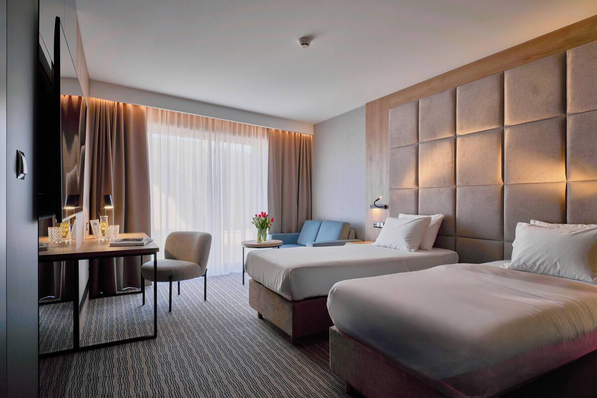 A hotel room with two beds, a desk, a chair, a TV, and a large window with curtains.