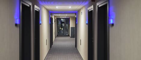A hotel corridor with a striped carpet, doors on both sides, and blue lighting on the ceiling.