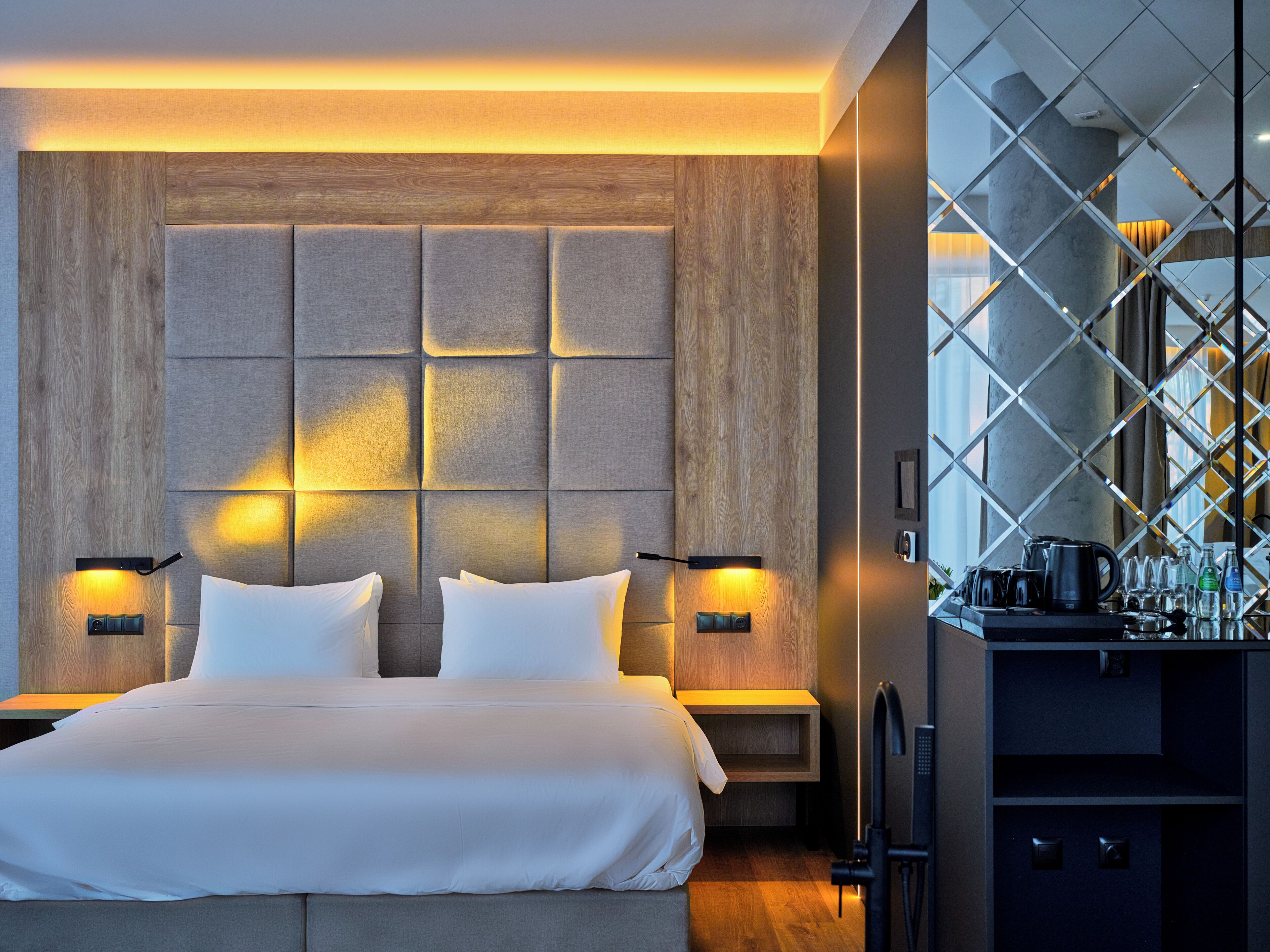 A modern hotel room with a large bed, two bedside tables, a built-in wardrobe, and a kitchen area with a coffee machine.