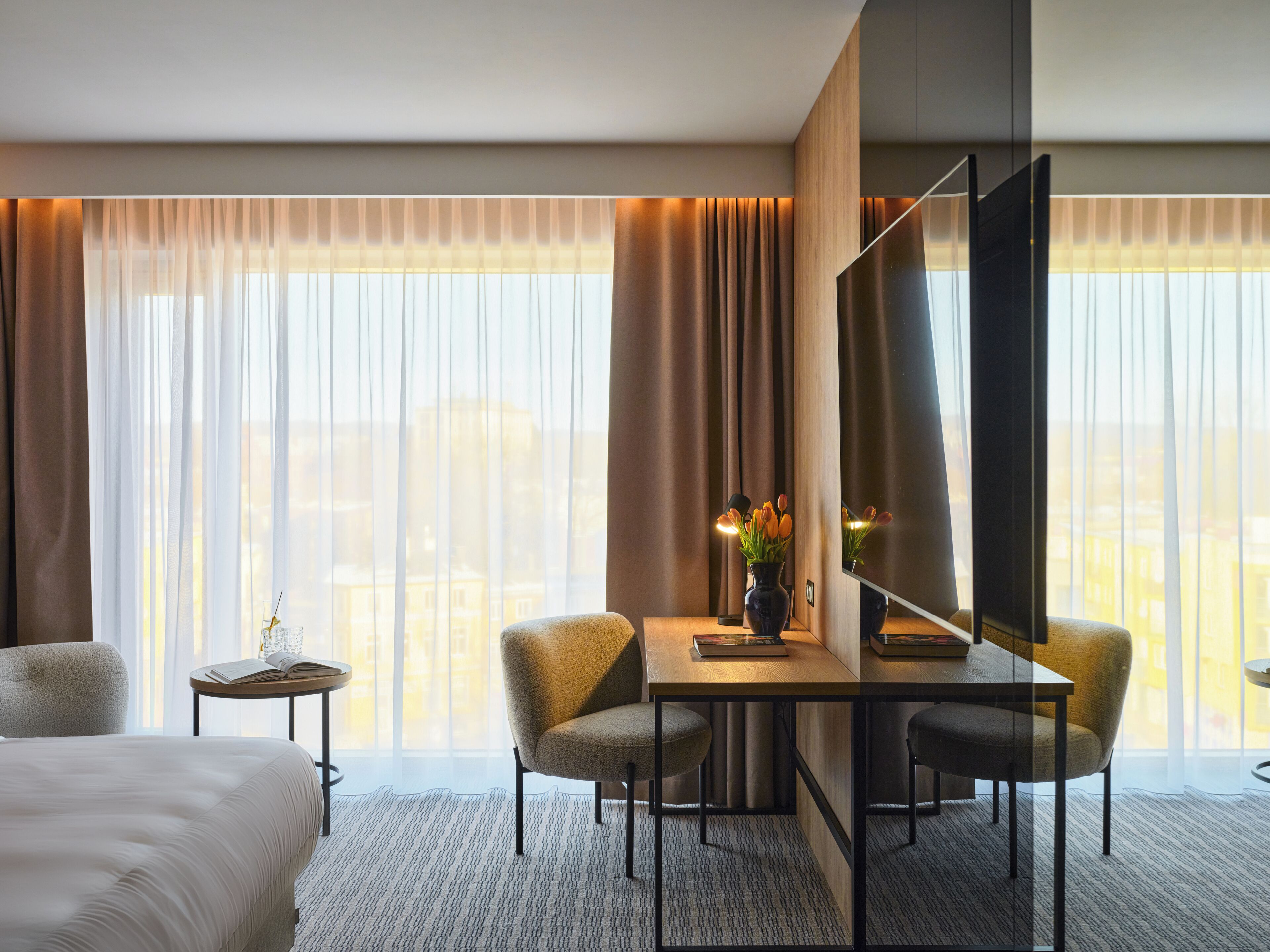 Habitación de hotel con cama, escritorio y dos sillas, televisión y un florero con flores.