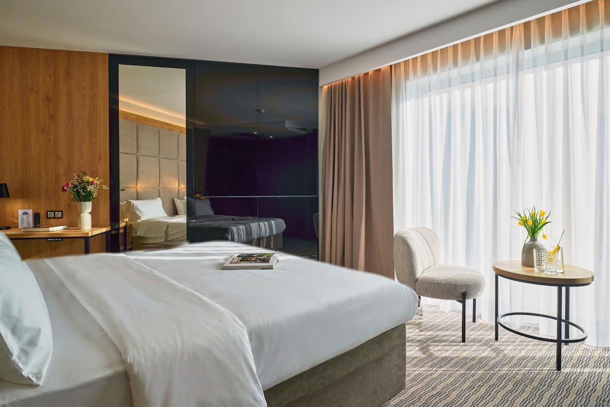 A modern hotel room with a large bed, a seating area with a chair, a small table with a vase, and a large window with curtains.