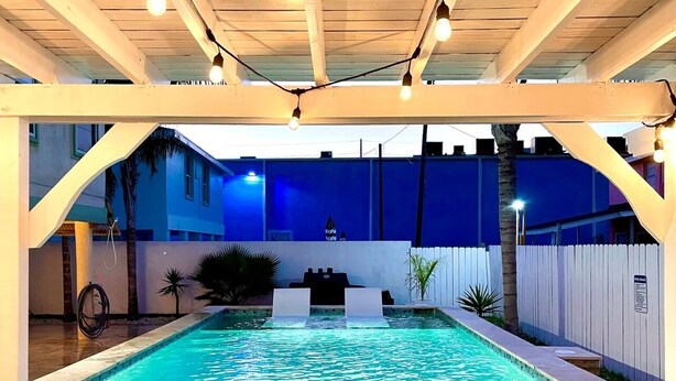 En swimmingpool med en kant av sten, under en pergola av trä med hängande lampor, omgiven av ett vitt staket och palmer.
