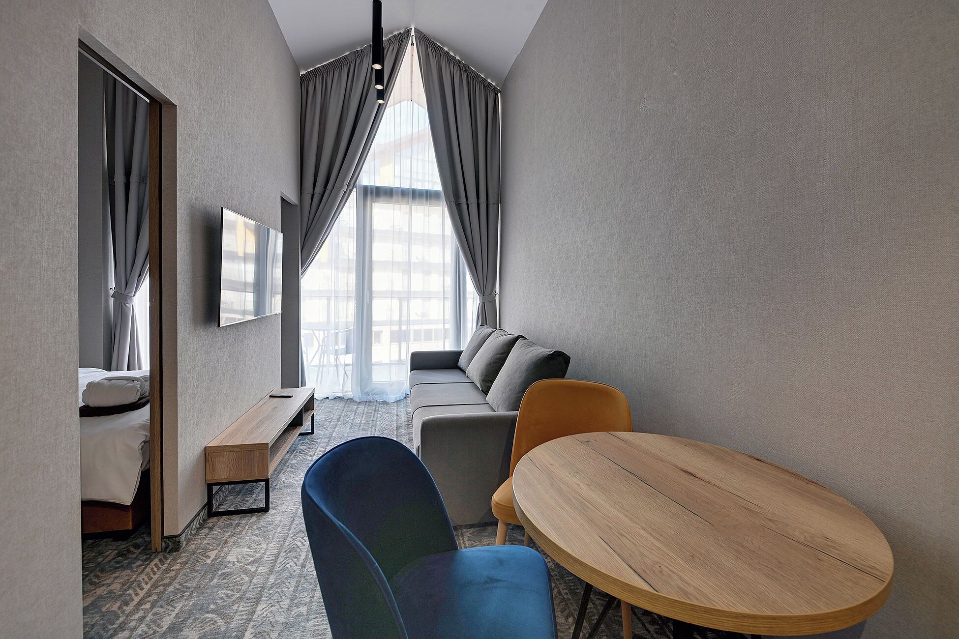 A modern hotel room with a sofa, a round wooden table, a blue chair, a TV mounted on the wall, and a large window with curtains.