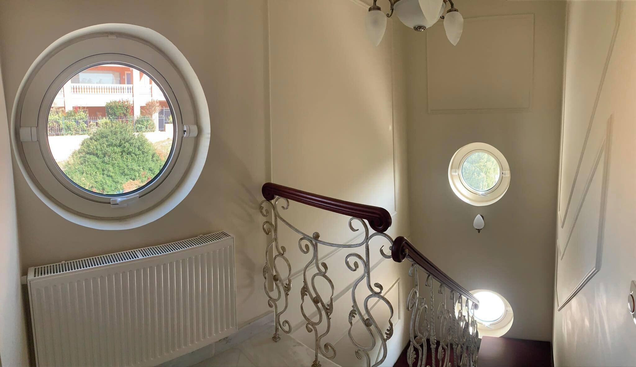 A staircase with ornate metal railings, a radiator, and two round windows offering views of greenery and a building.