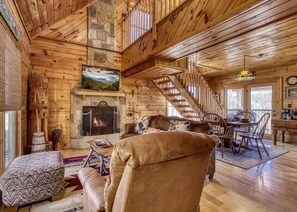 A cozy cabin interior with a stone fireplace, wooden walls, a dining area, and a staircase. - The Secluded Hideaway- Privacy, Romance, Hot Tub, & Pool Table (Sevierville)