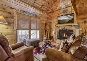 A cozy cabin interior with wooden walls, a stone fireplace, leather furniture, and a large window with a view. - The Secluded Hideaway- Privacy, Romance, Hot Tub, & Pool Table (Sevierville)