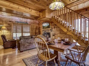 A wooden cabin interior with a dining table, chairs, a sofa, a fireplace, and a staircase. - The Secluded Hideaway- Privacy, Romance, Hot Tub, & Pool Table (Sevierville)
