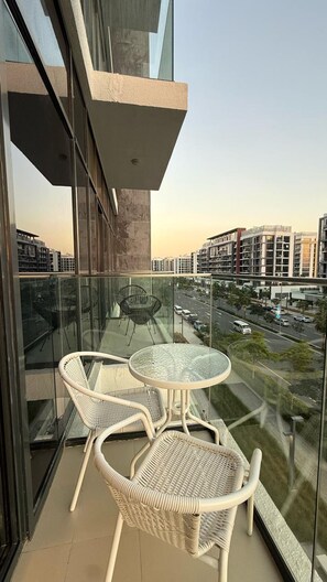 A balcony with wicker chairs and a glass table, overlooking a cityscape with modern buildings and a road. - Vesta - Luxury Studio - Riviera (Dubai)
