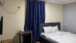 A hotel room with a bed, a bedside table with a phone, a small wooden side table, a blue curtain, and a wall-mounted air conditioner. - Kamson Hotel (Oba Ile)