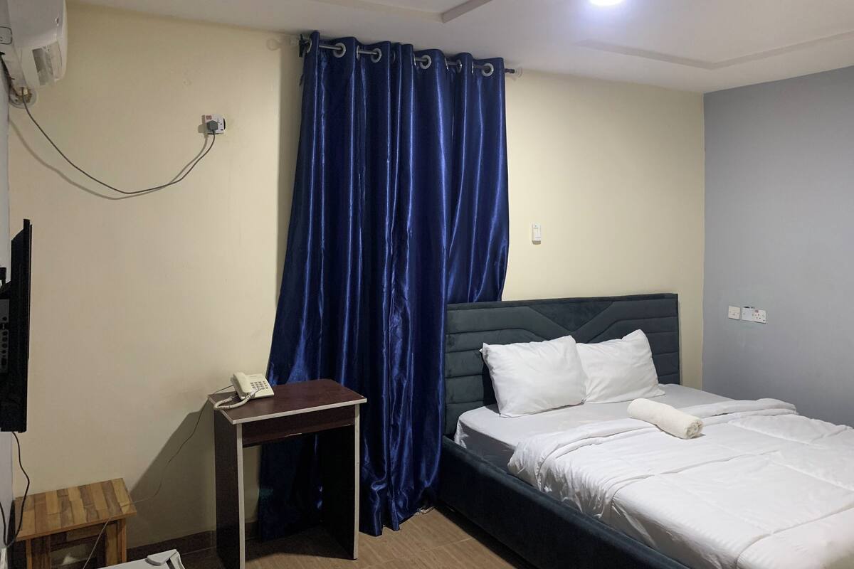 A hotel room with a bed, a bedside table with a phone, a small wooden side table, a blue curtain, and a wall-mounted air conditioner.