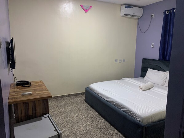 A hotel room with a bed, a wooden nightstand, an air conditioning unit, and a wall-mounted TV.