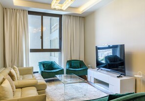 Ein modernes Wohnzimmer mit einer Couch, zwei Sesseln, einem Glastisch, einem Flachbildfernseher und einem großen Fenster mit Vorhängen.