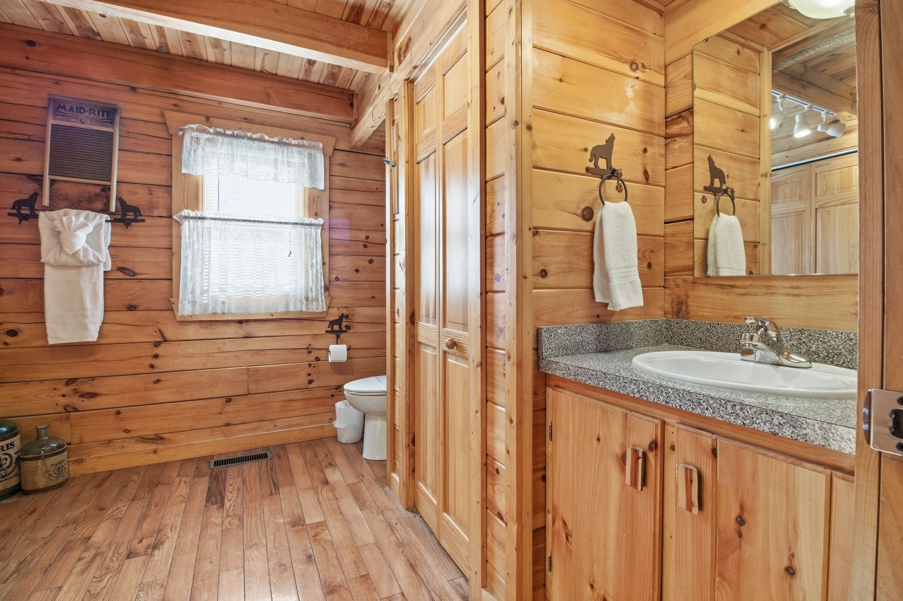 Une salle de bain de style rustique, avec des murs en bois, un plancher en bois, un lavabo doté d’un comptoir en granit, des toilettes et une fenêtre ornée de rideaux en dentelle.