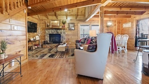 Interior acogedor de una cabaña de troncos con chimenea de piedra, pisos de madera, un sillón blanco y un comedor con mesa y sillas.