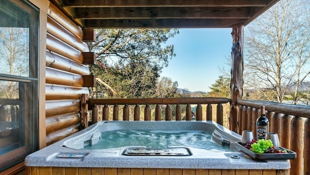 Un jacuzzi con terraza de madera, una cabaña de madera y una vista panorámica de árboles y montañas.