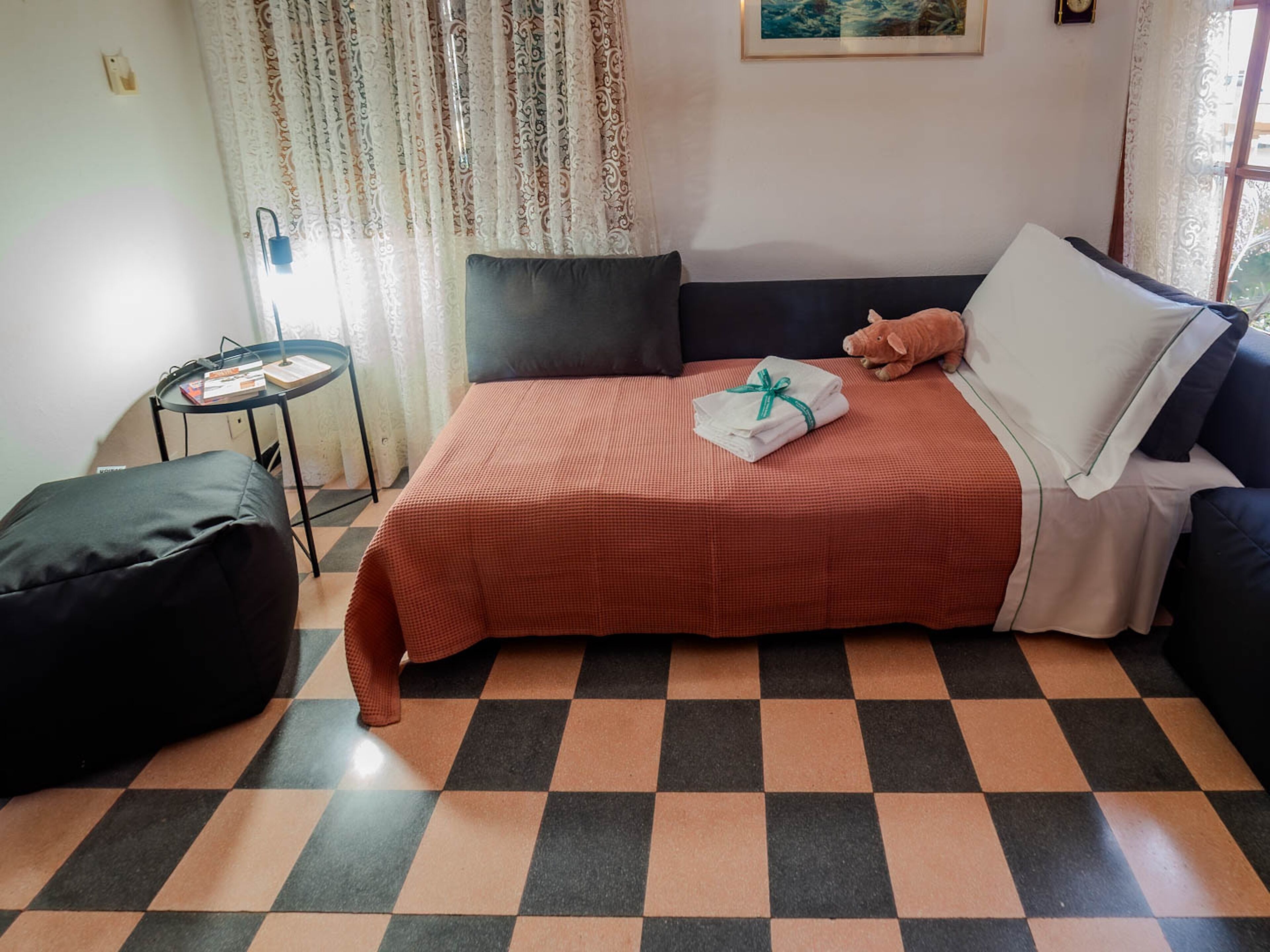 Une pièce avec un sol à carreaux, un lit recouvert d’une couverture rouge, une chaise noire, une petite table ronde sur laquelle sont posés des livres et une fenêtre avec des rideaux.