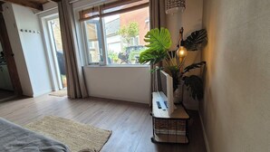 A cozy room with a wooden ceiling, a large window with curtains, a bedside table with a lamp, and a potted plant. - Cottage Strand & Centrum @NoordwijkMeer.nl (Noordwijk)