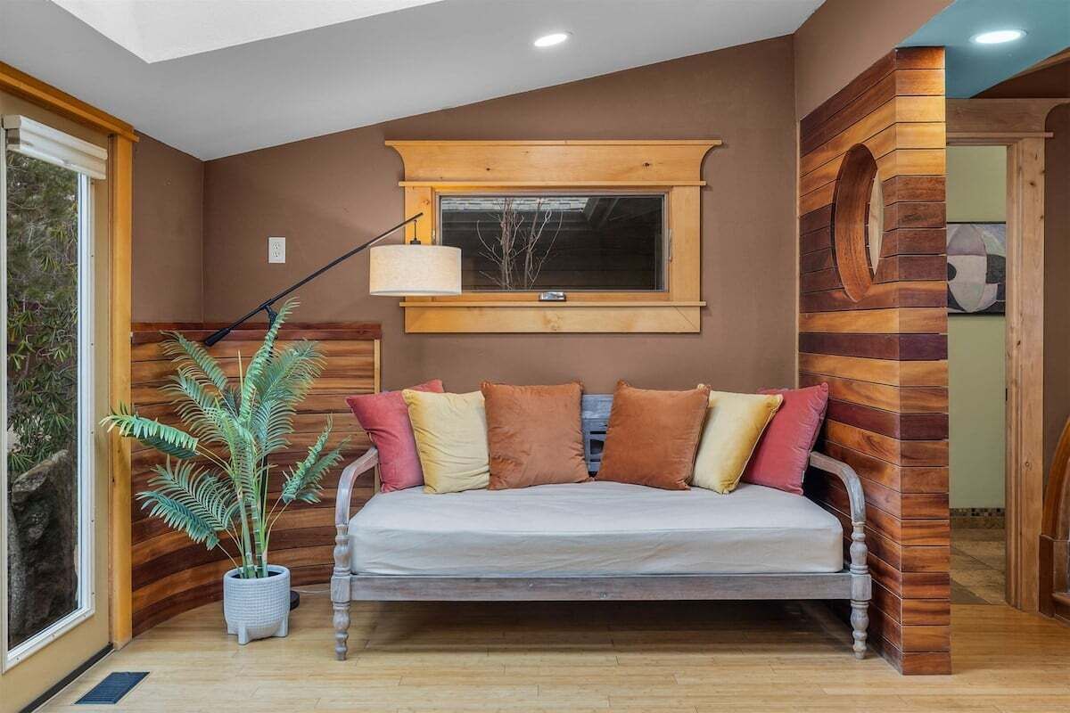 A cozy room with a wooden wall, a window with a view, a sofa with cushions, and a potted plant.