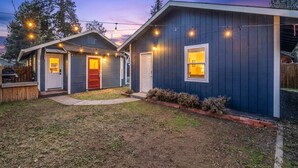 A two-story house with a dark blue exterior, a red door, and a small front yard with a wooden fence and a string of lights. - Bluebird Day - Pine Rosa Retreat, Cozy 2 BR, West side! (Bend)