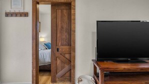 A room with a wooden door, a bed with blue bedding, a flat-screen TV on a wooden stand, and a wall-mounted coat rack with clips. - Bluebird Day - Pine Rosa Retreat, Cozy 2 BR, West side! (Bend)