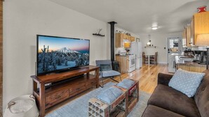 A modern living room with a television, wooden furniture, a brown sofa, and a kitchen in the background. - Bluebird Day - Pine Rosa Retreat, Cozy 2 BR, West side! (Bend)