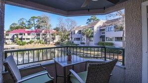 A balcony with wicker furniture, a table with a potted plant, and a view of buildings and trees across a body of water. - 7447 Anchorage: Pet Friendly 2 Bedroom Villa with Water Views, Sleeps 6 (Hilton Head Island)