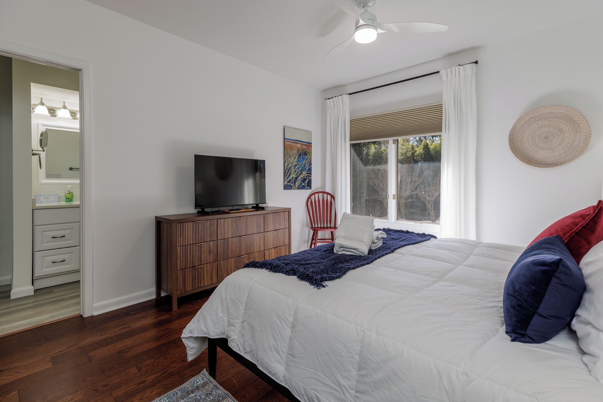 A bedroom with a bed, a television on a wooden dresser, a window with curtains, and a door leading to a bathroom.