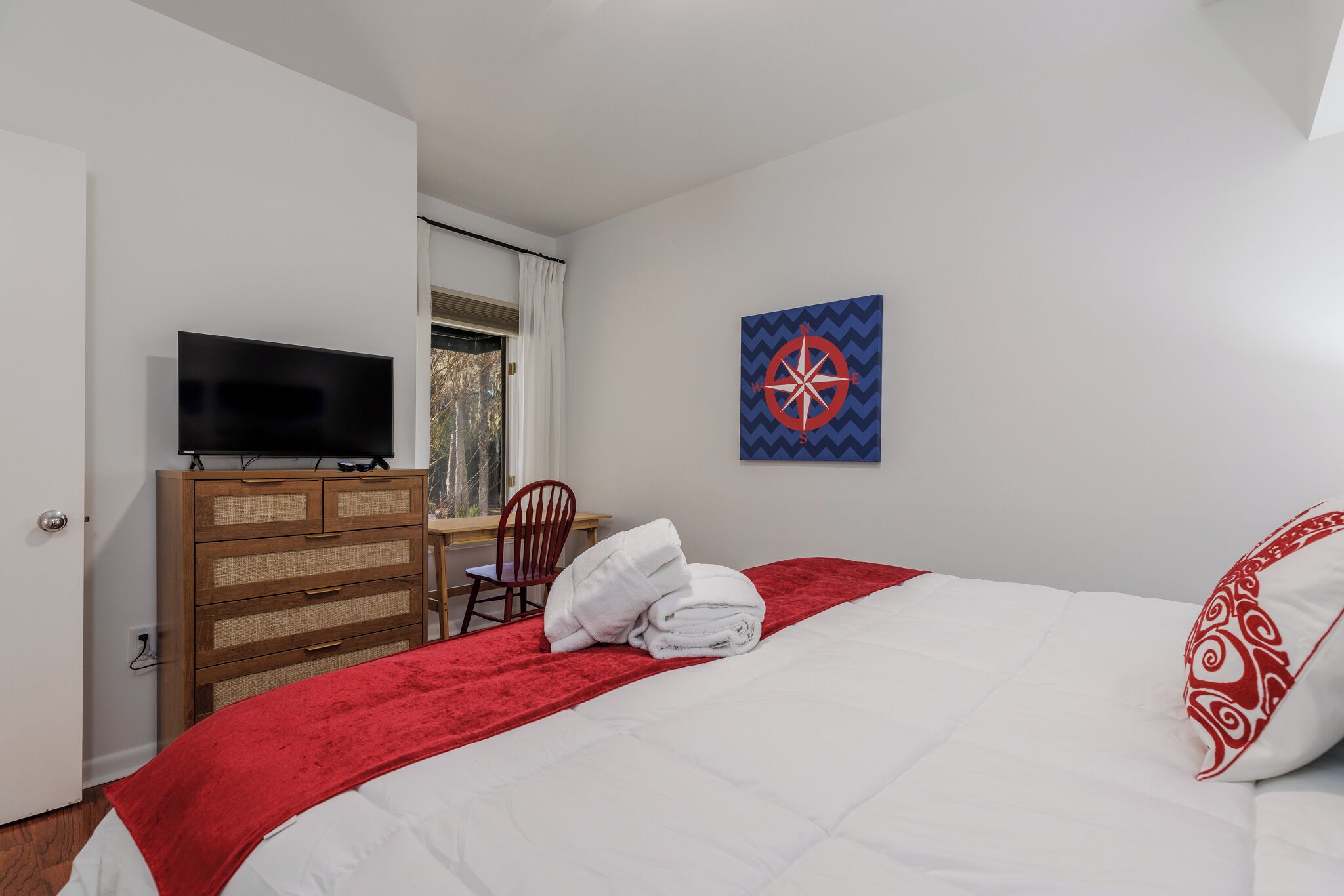 A hotel room with a bed, a TV on a wooden dresser, a red blanket, a white pillow, and a wall art piece.