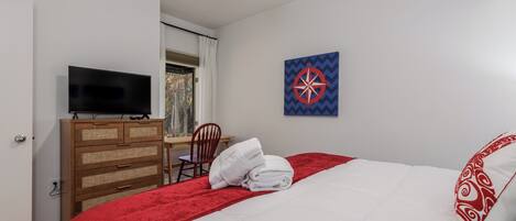 A hotel room with a bed, a TV on a wooden dresser, a red blanket, a white pillow, and a wall art piece.