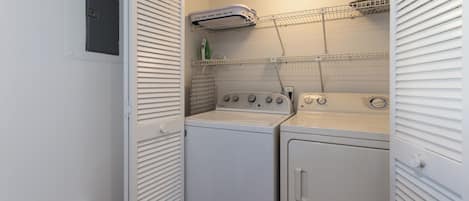 Uma lavanderia compacta com máquina de lavar e secadora, prateleiras acima para armazenamento e um exaustor no teto.