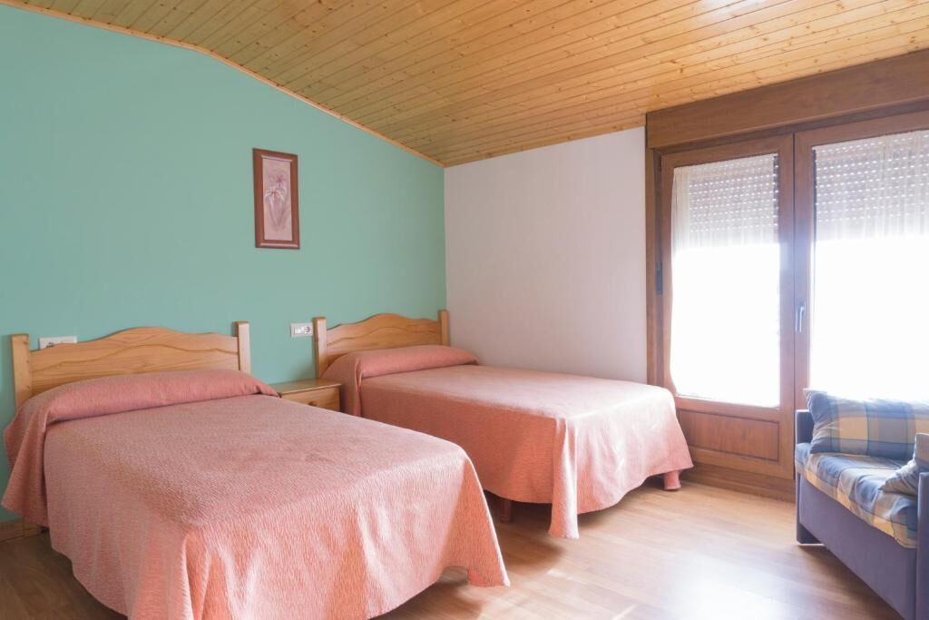 Una habitación con dos camas individuales, cabeceros de madera, techo de madera, una ventana con persianas y un sofá con funda a cuadros.