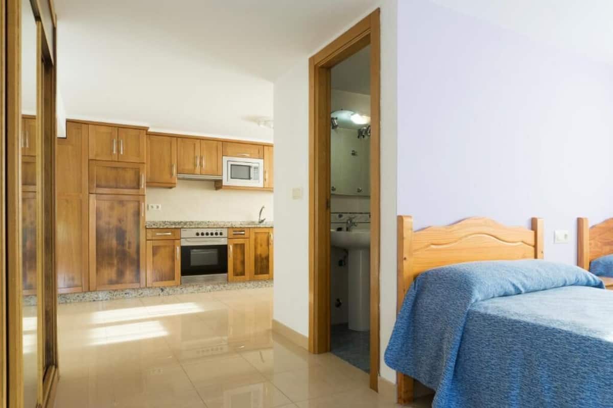 A room with a bed, a wooden headboard, and a view into a kitchen with wooden cabinets and a microwave.