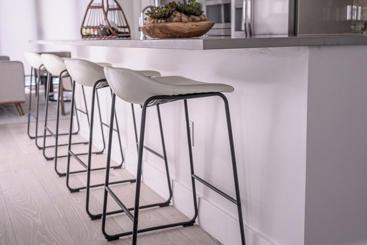 Une rangée de tabourets de bar avec des structures métalliques et des assises blanches, disposés le long d’un îlot de cuisine sur lequel se trouve un saladier en bois contenant des plantes.