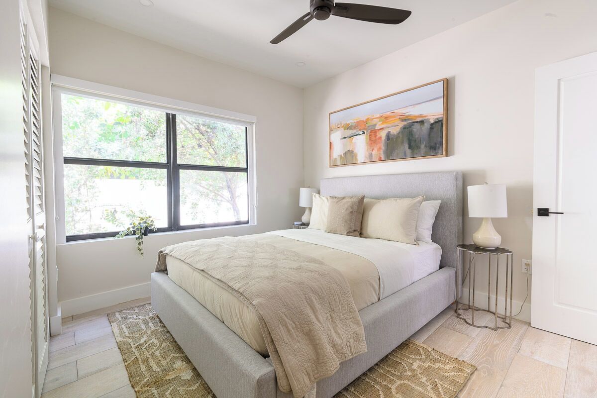 A bedroom with a bed, a ceiling fan, a window, a painting, and a bedside table.
