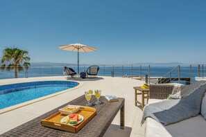 Ein Poolbereich mit Meerblick, eine Liege, ein Tisch mit Getränken und Snacks sowie ein Rattansofa.