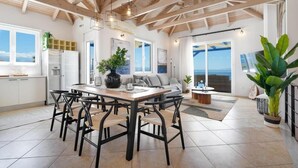 A spacious dining area with a wooden table and chairs, a large potted plant, and a view of the ocean through large windows. - Villa Hemera (Chania)